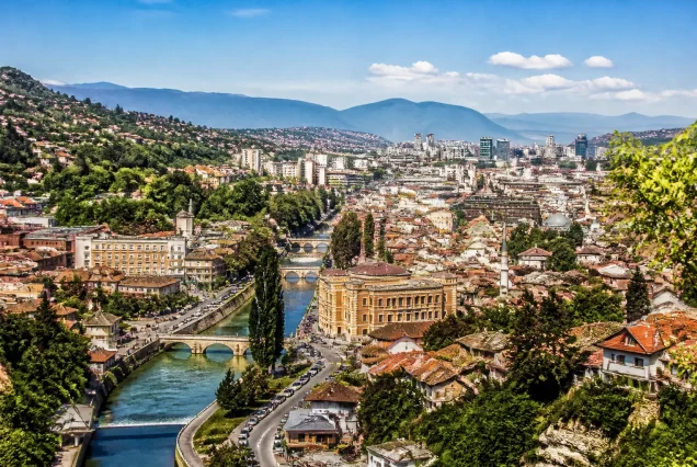 Saraybosna şehir merkezinde tarihi Osmanlı mimarisi, taş köprü ve arka planda yeşil dağlar